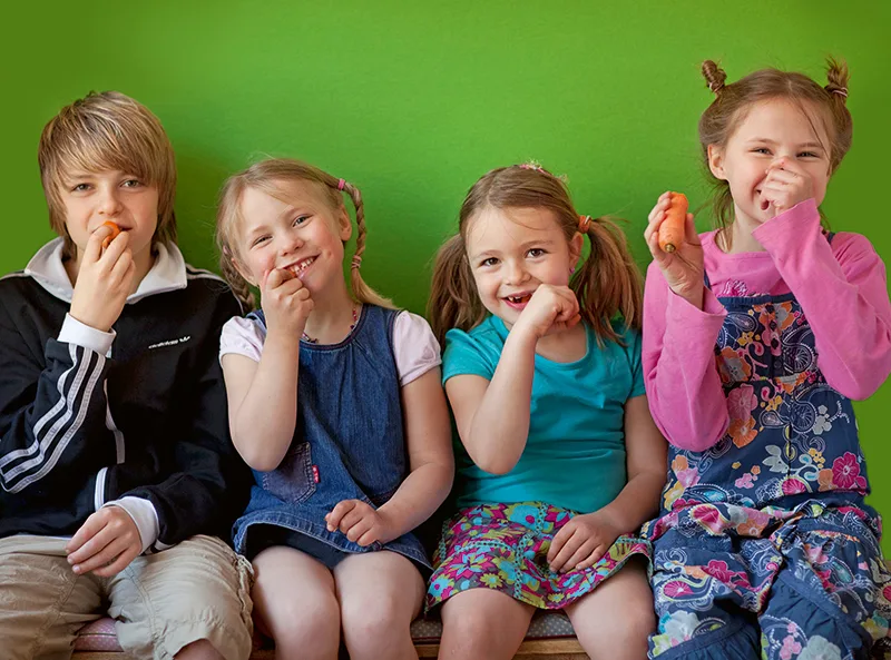 Kinder essen lachend Möhren – Titelmotiv des Magazins KOMPASS ERNÄHRUNG